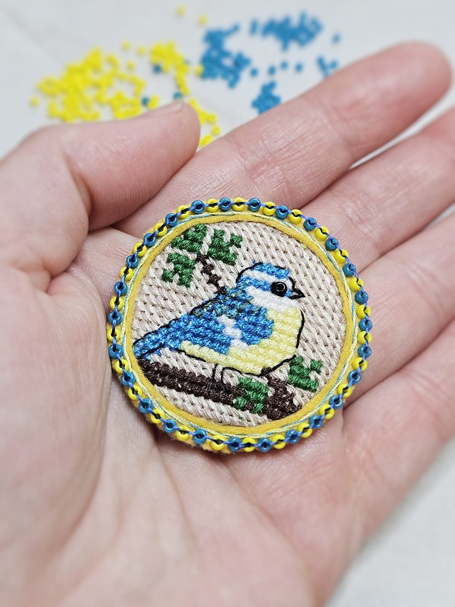Round cross-stitch brooch of a blue tit, edged with blue and yellow beads